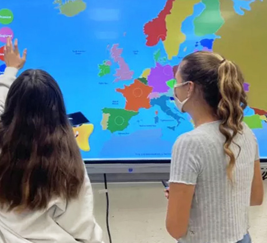Two students interacting with a digital map of Europe, one pointing at the screen while the other observes.